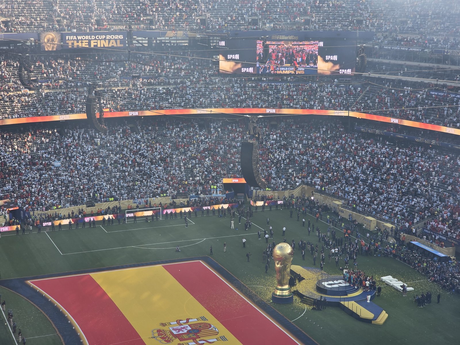 The FIFA World Cup 2026 trophy on the podium at MetLife Stadium, the Spain flag across the pitch and the stands packed beyond